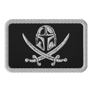 May include: A black and white embroidered patch featuring a Mandalorian helmet and crossed swords. The patch has a grey border.