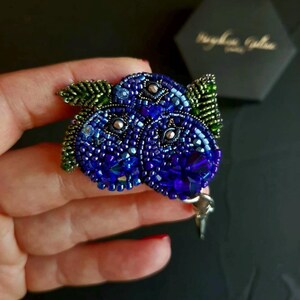 Könnte beinhalten: Handgefertigte Brosche in Form von Blaubeeren, mit aufwendiger Perlenarbeit in Blau- und Grüntönen. Die Brosche hat ein detailliertes Design mit einem Kristallakzent und einem Nadelverschluss. Ein einzigartiges Accessoire.