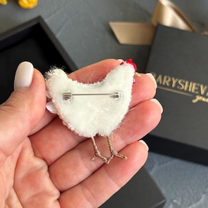 May include: A white chicken-shaped brooch with a red crest and gold-toned wire legs. The back of the brooch features a silver-coloured metal pin mechanism attached to a white felt backing, showcasing the construction of this handcrafted accessory.