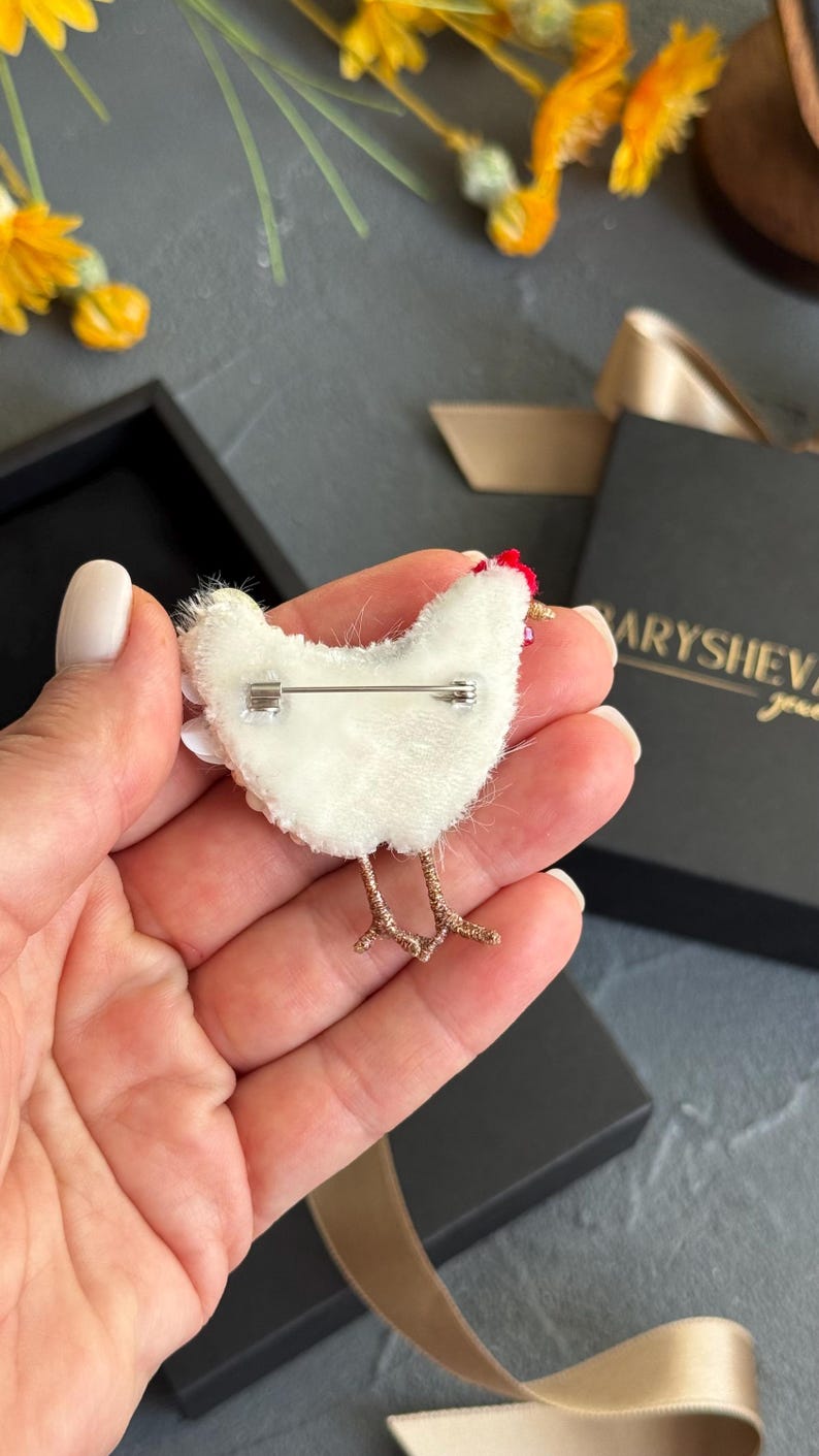 May include: A white chicken-shaped brooch with a red crest and gold-toned wire legs. The back of the brooch features a silver-coloured metal pin mechanism attached to a white felt backing, showcasing the construction of this handcrafted accessory.
