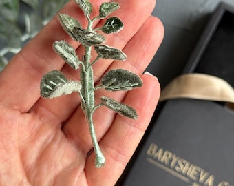 May include: A detailed, green embroidered brooch depicting a plant stem with leaves. The leaves have a soft, textured finish. The brooch is held in a hand, with a black box and the text "BARYSHEVA GAL" visible in the background.