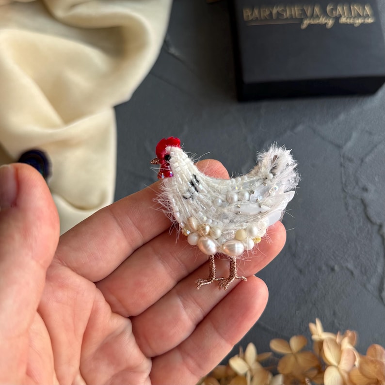 May include: A handcrafted chicken brooch featuring white thread, iridescent beads, and lustrous pearls. The bird has a small red bead for a comb and thin, metallic wire legs. The design is textured with fine fibres and various white embellishments.