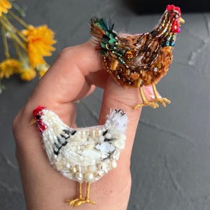 May include: Two chicken-shaped brooches crafted with intricate beadwork. One brooch features brown, amber, and green beads with a red comb. The second brooch is made of white beads, pearls, and clear crystals with a red comb and gold-toned metal feet.