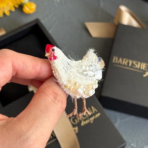 May include: A white chicken brooch crafted with intricate beadwork, featuring a bright red comb and wattle, textured white feathers made of small beads and pearls, and thin metallic copper-coloured legs extending from the bottom of the body.