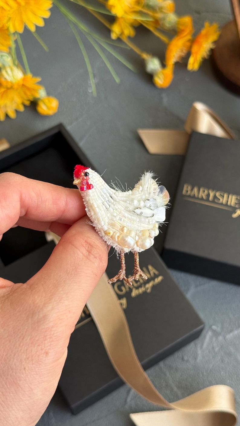May include: A white chicken brooch crafted with intricate beadwork, featuring a bright red comb and wattle, textured white feathers made of small beads and pearls, and thin metallic copper-coloured legs extending from the bottom of the body.