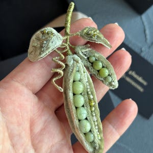May include: A handcrafted pea pod brooch. The design features a light green velvet pod, filled with round, pale green beads. The leaves and stem are also light green, with intricate bead detailing. A unique, nature-inspired accessory.