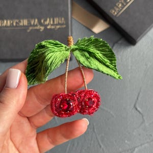 May include: A handmade cherry brooch featuring red beaded cherries and green velvet leaves. The stems are gold-coloured wire. The brooch is held in a hand, with a blurred background.