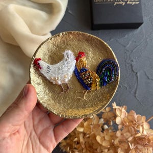 May include: A round, textured gold-toned dish featuring two embroidered bird pins. One pin is a white chicken with pearl-like accents, and the other is a rooster with multi-coloured beads in shades of blue, yellow, and green with metallic sequins.
