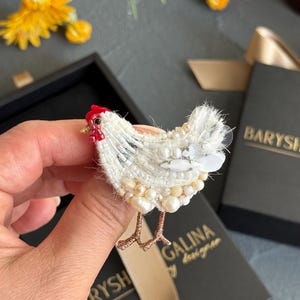 May include: A chicken-shaped brooch crafted with white beads, sequins, and pearls. The bird features a small red beaded comb, a tiny black eye, and thin metallic gold-toned legs. The textured surface is composed of layered white embroidery and beads.