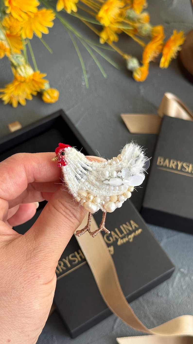May include: A chicken-shaped brooch crafted with white beads, sequins, and pearls. The bird features a small red beaded comb, a tiny black eye, and thin metallic gold-toned legs. The textured surface is composed of layered white embroidery and beads.