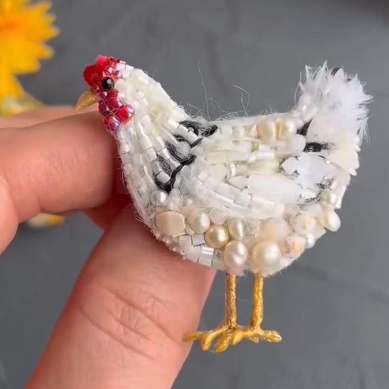 May include: A handcrafted chicken brooch, featuring a white beaded body with black accents and pearl embellishments. The chicken has a red comb and wattle, with gold-colored legs. A unique, handmade accessory.