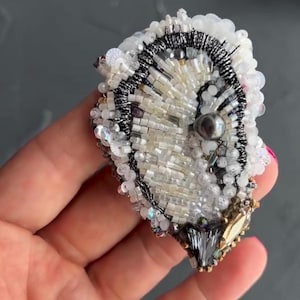 Op de afbeelding: Een handgemaakte, sierlijke broche met een centrale parel en ingewikkeld kralenwerk. De broche heeft witte, zilveren en zwarte kralen, met een donkergrijze rand en een mix van pailletten. De onderkant heeft een goud-grijze kristallen accent.