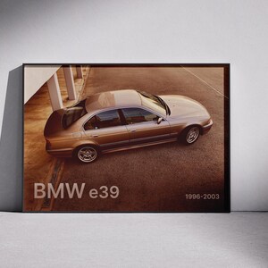 May include: A framed print featuring a gold-colored BMW e39 sedan from 1996-2003. The car is parked on a brown surface, with the text "BMW e39" and the production years displayed in white.