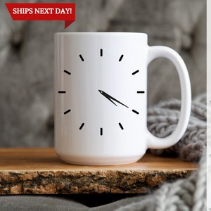 May include: White ceramic mug with a black clock face design. The clock face shows the time as 1:00. The mug has a black handle. The mug is sitting on a wooden surface.