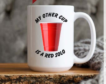 May include: White ceramic mug with a red Solo cup graphic and the text "MY OTHER CUP IS A RED SOLO". The mug is on a wooden surface. The mug is a novelty item.