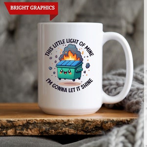 May include: White ceramic coffee mug with a green, yellow, and black cartoon image of a dumpster on fire. The text "This little light of mine I'm gonna let it shine" is printed on the mug.