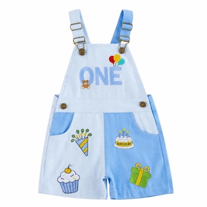 May include: Light blue and medium blue colour-blocked dungarees featuring adjustable metal buckles. The chest displays the word ONE with a small teddy bear and three balloons. The shorts feature embroidered party motifs including a cupcake, gift.