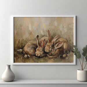 May include: A framed print depicting three brown rabbits nestled together in a field of long grass. The artwork uses a soft, muted colour palette, highlighting the rabbits' fur and long ears. The print is presented in a white frame.
