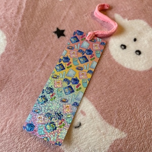 May include: A holographic bookmark with a retro video game design, including consoles and controllers in a repeating pattern. It has a pink cord attached. The bookmark is set against a pink patterned background.