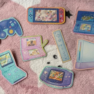 May include: A selection of colourful stickers depicting different gaming consoles and controllers. Includes a blue GameCube controller, a Nintendo Switch, a Nintendo DS, a Wii remote, a Game Boy Advance, and a Wii console.
