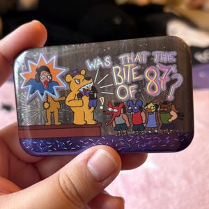 May include: A rectangular pin with a cartoon design. The pin features a yellow bear character with a purple hat, a person in a blue shirt, and the text "WAS THAT THE BITE OF 87?". Other cartoon characters are also present.