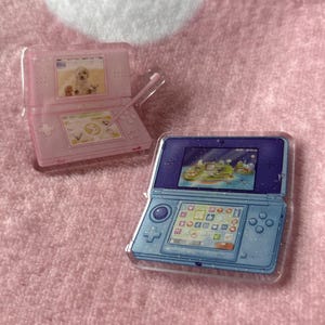 May include: Two clear acrylic game console charms. One is pink with a dog image on the screen, the other is blue with a landscape image. Both have glitter accents and are open, showing the screens and buttons.