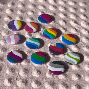 May include: A collection of 12 round button badges featuring various pride flags, including the bisexual, transgender, lesbian, gay, pansexual, and asexual flags. The badges are all white with a colourful design.