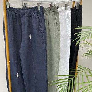 May include: Four pairs of linen trousers hanging on a gold rack. The trousers are in navy blue, sage green, white, and black.