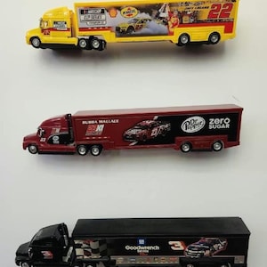May include: Three toy semi-trailer trucks with different paint schemes and sponsors. The first truck is yellow with a Pennzoil logo and the number 22. The second truck is red with a Dr. Pepper logo and the number 23. The third truck is black with a Goodwrench logo and the number 3.