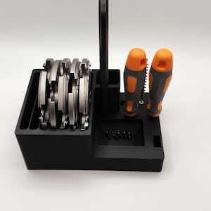 Puede incluir: Organizador de plástico negro con asa, que contiene varias hojas de sierra circular plateadas y dos destornilladores naranja y grises.