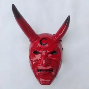 May include: A glossy red mask with two upward-curving horns, each tipped with black. The mask features a stern expression, eye holes, and a crescent moon symbol above the eyes. The mask is likely for costume or decorative purposes.
