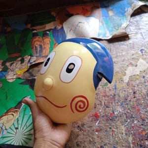 Può includere: Una maschera di personaggio dei cartoni animati gialla e blu con grandi occhi, un naso piccolo e un design a spirale rossa sulla guancia. La maschera ha un design per capelli blu ed è tenuta in una mano.