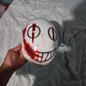 May include: White mask with black eye outlines, a red vertical line, and a red slash across the cheek. The mask has a wide, black-lined grin. The mask is held in a hand.