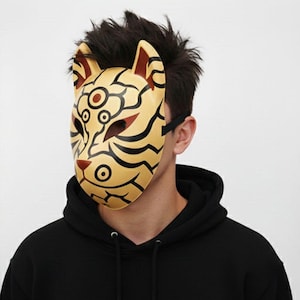 Può includere: Una maschera da volpe beige e nera con dettagli rossi. La maschera presenta un design dettagliato con strisce nere e motivi circolari. Presenta sporgenze simili a orecchie ed è tenuta in posizione da un cinturino nero.