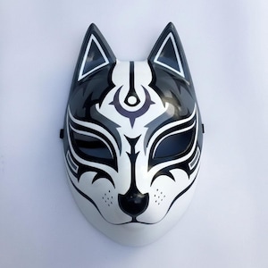 Könnte beinhalten: Eine weiße, schwarze und graue Fuchmaske mit einem symmetrischen Design. Die Maske hat spitze Ohren, dunkle Augenöffnungen und komplizierte Muster um die Augen und die Nase. Die Maske hat eine glänzende Oberfläche.