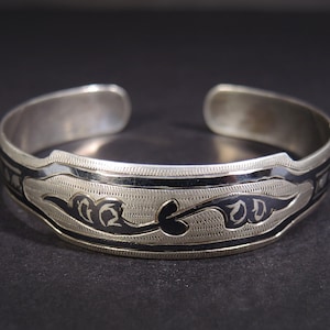 May include: Silver cuff bracelet with a detailed engraved design. The bracelet features a central band with a black floral pattern and a textured background. The edges are bordered with black lines. A stylish piece of jewelry.