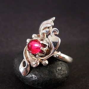 May include: A silver ring with a bright red gemstone. The ring features an ornate, floral design with curved, leaf-like elements. The gemstone is round and set in the center of the design.