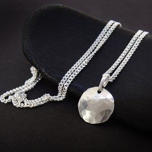 Puede incluir: Un collar de plata con un colgante redondo martillado. La cadena es de estilo curb.