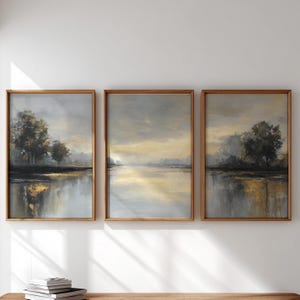May include: Set of three framed landscape paintings. Each artwork depicts a serene waterscape with trees and a reflective surface. The color palette includes muted tones of gray, gold, and brown, creating a tranquil atmosphere. The frames are made of wood.