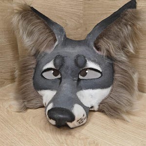 May include: A grey and white wolf mask with large, upright ears and fluffy, brown fur around the face. The mask has a detailed painted design with expressive eyes and a black nose, suitable for cosplay or costume use.