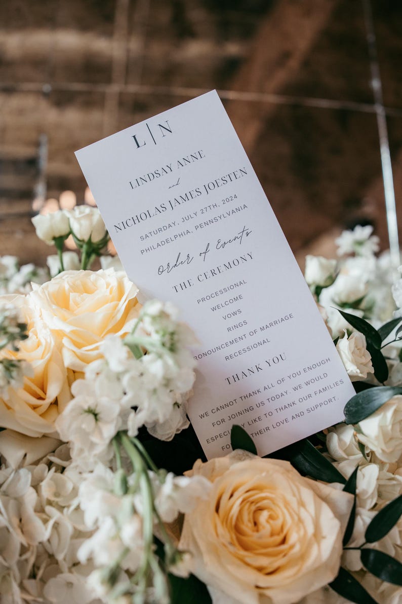 Pode incluir: Um programa de casamento com os nomes de Lindsay Anne e Nicholas James Joesten, e a data de 27 de julho de 2024. O programa &eacute; rodeado por rosas brancas e p&ecirc;ssego e outras flores brancas.