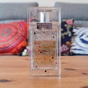 Könnte beinhalten: Ein rechteckiger Parfümflakon in einer transparenten, dekorativen Hülle. Die Flasche hat einen silbernen Deckel und ist mit einer hellen, bernsteinfarbenen Flüssigkeit gefüllt. Das Gehäuse ist mit roten und klaren Kristallen und dem Text "LET THIS BE PARTY" verziert.
