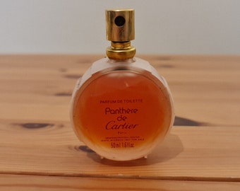 Può includere: Un flacone di profumo Panthère de Cartier in vetro trasparente. Il flacone è rotondo e ha un tappo dorato. L'etichetta è rossa e bianca e dice "Parfum de Toilette Panthère de Cartier Paris". Il flacone contiene 50 ml o 1,6 fl oz.