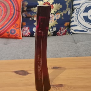 Könnte beinhalten: Eine hochgewachsene, geschwungene Parfümflasche in dunklem Rot. Die Flasche hat eine rechteckige Form mit abgerundeten Kanten und einem dunklen Verschluss. Der Markenname und der Produktname sind auf der Vorderseite aufgedruckt. Die Flasche steht auf einer Holzoberfläche.
