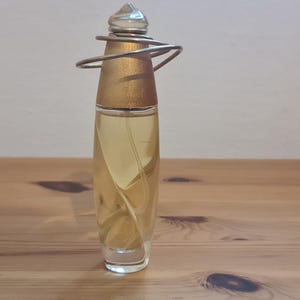 Puede incluir: Un frasco de perfume de vidrio transparente y dorado con un diseño de alambre plateado alrededor de la parte superior. El frasco está sobre una superficie de madera.