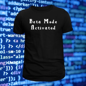 May include: A black short-sleeved t-shirt featuring the white pixelated text "Beta Mode Activated" centered on the chest. The garment has a classic crew neckline and a straight hem, presenting a simple design with a clear, bold message in a retro font.