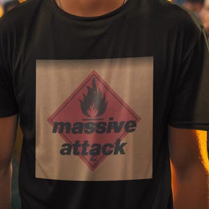Puede incluir: Una camiseta negra con un diseño gráfico que presenta un rombo rojo, una llama negra y las palabras "massive attack". La camiseta la lleva una persona entre una multitud.