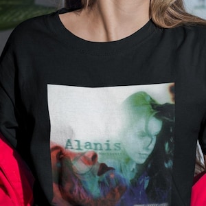 May include: Black t-shirt featuring a graphic print of the album cover for "Jagged Little Pill" by Alanis Morissette. The album cover depicts a woman with long dark hair against a blurred background.