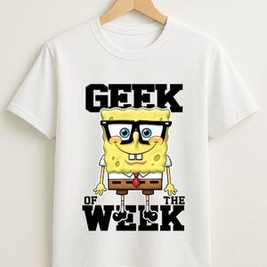 May include: White t-shirt featuring a cartoon image of SpongeBob SquarePants wearing glasses and a tie. The text "GEEK OF THE WEEK" is printed above and below the character in black block letters. The shirt is hanging on a wooden hanger.