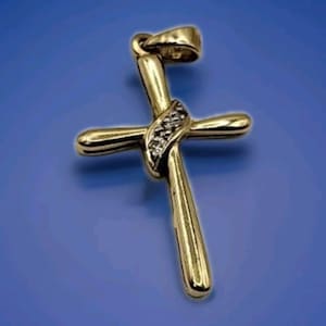 May include: A gold-colored cross pendant with a small loop at the top for a chain. The cross has a curved design with a band of small, clear stones at the intersection. The background is a solid blue.